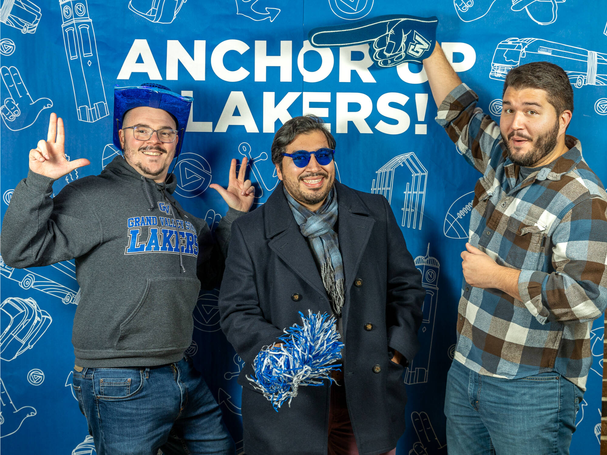 three individuals, one on the left holding up the anchor up finger sign wearing a blue cowboy hat, one in the middle holding up a pompom, one on the right pointing a foam finger at the individual on the left
